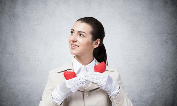 Smiling young woman holding retro red phone. Call center operator in white business suit posing with landline phone in hand. Hotline telemarketing concept. Business assistance and consultation.