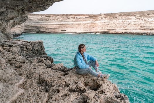 A woman in a blue jacket sits on a rock above a cliff above the sea, looking at the stormy ocean. Girl traveler rests, thinks, dreams, enjoys nature. Peace and calm landscape, windy weather