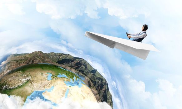 Pilot in leather helmet and goggles driving paper plane on background of blue cloudy sky. Traveling around the world. Funny man flying in small paper airplane. Earth horizon with high mountain range