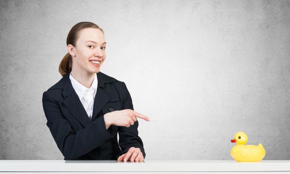 Business woman sits at a white table. Points finger at a yellow plastic toy duck