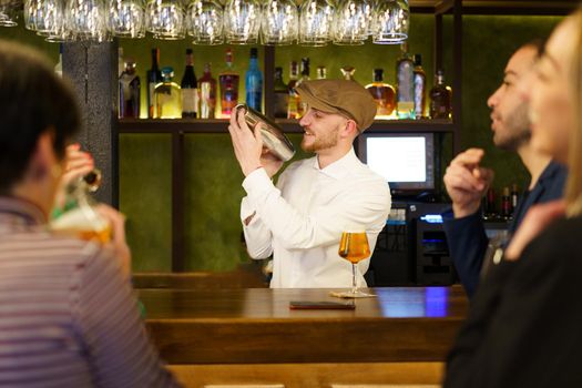 Glad bearded man, in white shirt and cap, smiling and mixing cocktail ingredients in shaker, while standing behind counter near male and female customers in bar