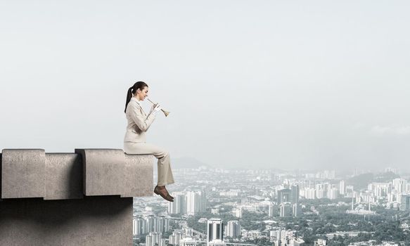 Elegant young woman playing trumpet on edge of roof. Girl in white business suit and gloves with music brass instrument posing on roof of building. Blue sky and modern metropolis with skyscrapers.