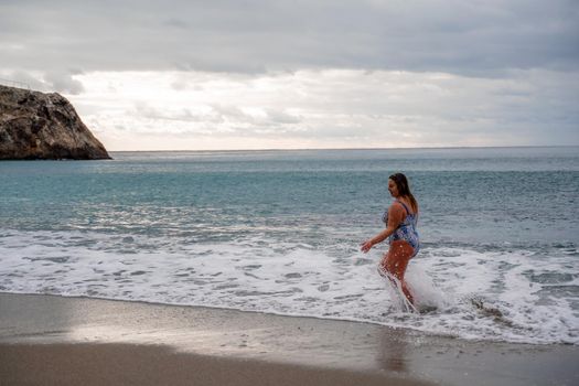 A plump woman in a bathing suit enters the water during the surf. Alone on the beach, Gray sky in the clouds, swimming in winter