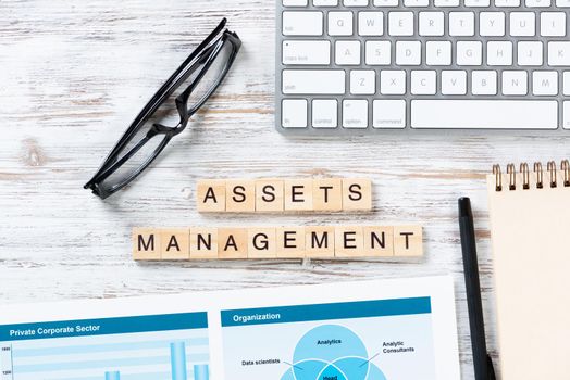 Asset management concept with letters on cubes. Still life of office workplace with supplies. Flat lay vintage wooden desk with computer keyboard and analytic report. Capital investment company