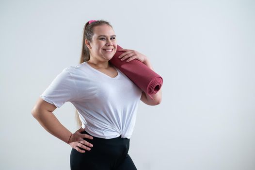 Young fat caucasian woman holding a sport mat. Charming plus size model in sportswear stands on a white background.