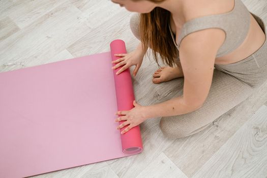 A young woman rolls a pink fitness or yoga mat before or after exercising, exercising at home in the living room or in a yoga studio. Healthy habits, keep fit, weight loss concept. Closeup photo.