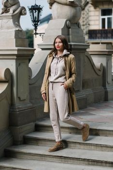 Portrait of fashionable women in beige sports suit, trench coat and stylish suede loafer posing on the stone stairs. street look fashion