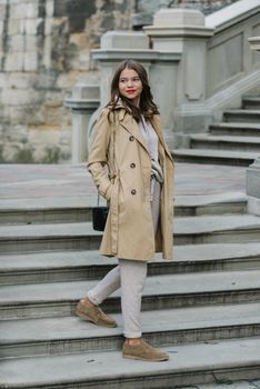 Portrait of fashionable women in beige sports suit, trench coat and stylish suede loafer posing on the stone stairs. street look fashion