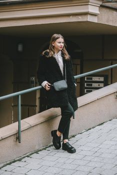 Fashion photo of young woman in black fur coat, jeanse and shoes at city street. beige jacket with a hood