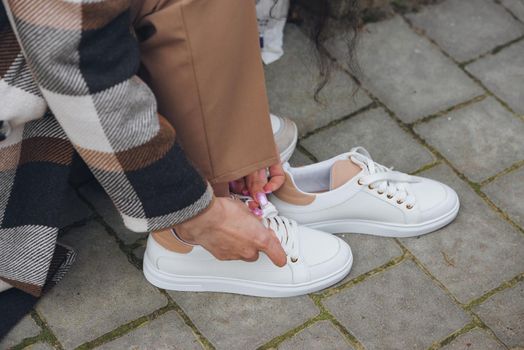 Closeup photo of young woman wearing checkered long coat, and beige pants . Lady posing on city street. Women hands ties laces on sneakers