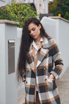 Closeup portrait of young beautiful fashionable woman wearing checkered long coat, beige pants and white blouse . Lady posing on city street.. Female fashion.