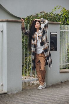 Closeup portrait of young beautiful fashionable woman wearing checkered long coat, beige pants and white blouse . Lady posing on city street.. Female fashion.