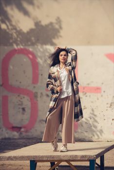 Closeup portrait of young beautiful fashionable woman wearing checkered long coat, beige pants and white blouse . Lady posing on city building background. Female fashion.