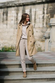 Portrait of fashionable women in beige sports suit, trench coat and stylish suede loafer posing on the stone stairs. street look fashion