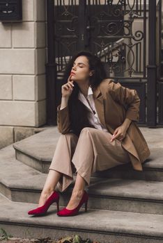 Close up portrait of young beautiful woman with long brunette curly hair posing against building background. woman wearing a white blouse and beige pants and a jacket