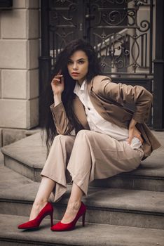 Close up portrait of young beautiful woman with long brunette curly hair posing against building background. woman wearing a white blouse and beige pants and a jacket