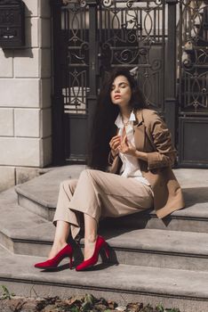 Close up portrait of young beautiful woman with long brunette curly hair posing against building background. woman wearing a white blouse and beige pants and a jacket