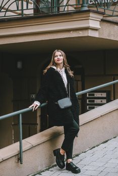 Fashion photo of young woman in black fur coat, jeanse and shoes at city street. beige jacket with a hood