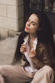 Close up portrait of young beautiful woman with long brunette curly hair posing against building background. woman wearing a white blouse and beige pants and a jacket