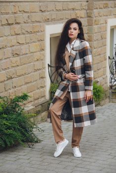 Closeup portrait of young beautiful fashionable woman wearing checkered long coat, beige pants and white blouse . Lady posing on city street.. Female fashion.