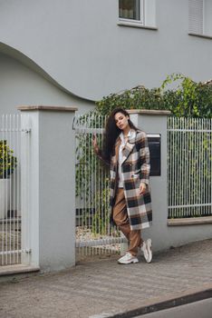 Closeup portrait of young beautiful fashionable woman wearing checkered long coat, beige pants and white blouse . Lady posing on city street.. Female fashion.