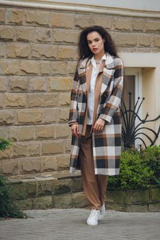 Closeup portrait of young beautiful fashionable woman wearing checkered long coat, beige pants and white blouse . Lady posing on city street.. Female fashion.