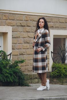 Closeup portrait of young beautiful fashionable woman wearing checkered long coat, beige pants and white blouse . Lady posing on city street.. Female fashion.