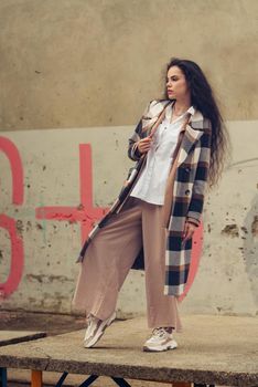 Closeup portrait of young beautiful fashionable woman wearing checkered long coat, beige pants and white blouse . Lady posing on city building background. Female fashion.