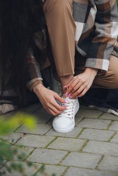 Closeup photo of young woman wearing checkered long coat, and beige pants . Lady posing on city street. Women hands ties laces on sneakers