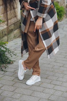 Closeup photo of beautiful fashionable woman wearing checkered long coat, beige pants, white blouse and sneakers. Lady posing on city street.