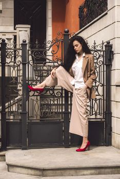 Close up portrait of young beautiful woman with long brunette curly hair posing against building background. woman wearing a white blouse and beige pants and a jacket