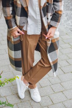 Closeup photo of beautiful fashionable woman wearing checkered long coat, beige pants, white blouse and sneakers. Lady posing on city street.