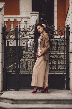 Close up portrait of young beautiful woman with long brunette curly hair posing against building background. woman wearing a white blouse and beige pants and a jacket