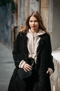 Fashion photo of young woman in black fur coat, jeanse and shoes at city street. beige jacket with a hood