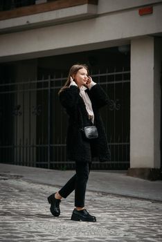 Fashion photo of young woman in black fur coat, jeanse and shoes at city street. beige jacket with a hood