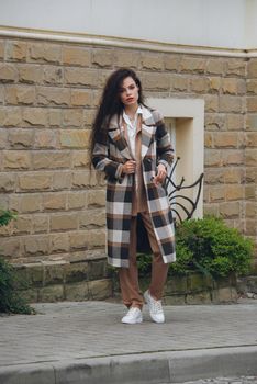 Closeup portrait of young beautiful fashionable woman wearing checkered long coat, beige pants and white blouse . Lady posing on city street.. Female fashion.