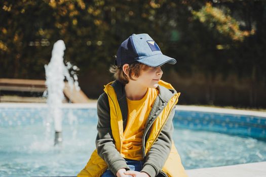 Schoolboy sitting on a fountain in public park during spring sunny day thoughtfully looking aside. Kid in yellow vest chilling in park with water sprinkling in the background. Summer holidays outside.