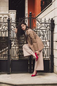 Close up portrait of young beautiful woman with long brunette curly hair posing against building background. woman wearing a white blouse and beige pants and a jacket