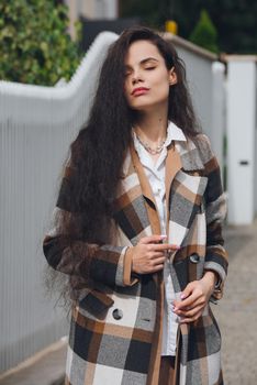 Closeup portrait of young beautiful fashionable woman wearing checkered long coat, beige pants and white blouse . Lady posing on city street.. Female fashion.