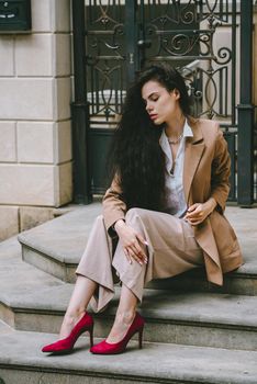 Close up portrait of young beautiful woman with long brunette curly hair posing against building background. woman wearing a white blouse and beige pants and a jacket