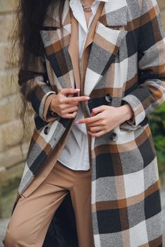 Closeup portrait of young beautiful fashionable woman wearing checkered long coat, beige pants and white blouse . Lady posing on city street.. Female fashion.