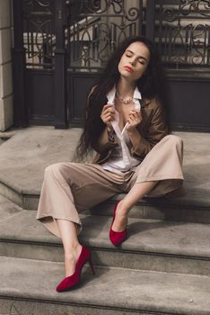 Close up portrait of young beautiful woman with long brunette curly hair posing against building background. woman wearing a white blouse and beige pants and a jacket