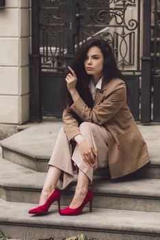 Close up portrait of young beautiful woman with long brunette curly hair posing against building background. woman wearing a white blouse and beige pants and a jacket