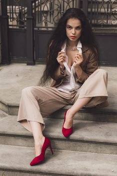 Close up portrait of young beautiful woman with long brunette curly hair posing against building background. woman wearing a white blouse and beige pants and a jacket