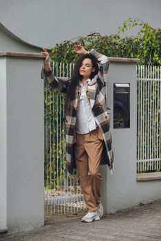 Closeup portrait of young beautiful fashionable woman wearing checkered long coat, beige pants and white blouse . Lady posing on city street.. Female fashion.