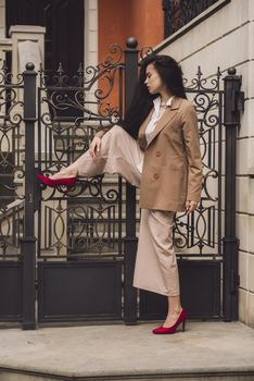 Close up portrait of young beautiful woman with long brunette curly hair posing against building background. woman wearing a white blouse and beige pants and a jacket