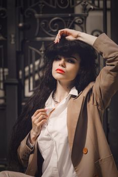 Close up portrait of young beautiful woman with long brunette curly hair posing against building background. woman wearing a white blouse and beige jacket