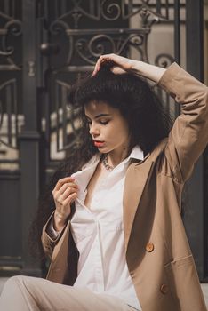 Close up portrait of young beautiful woman with long brunette curly hair posing against building background. woman wearing a white blouse and beige jacket