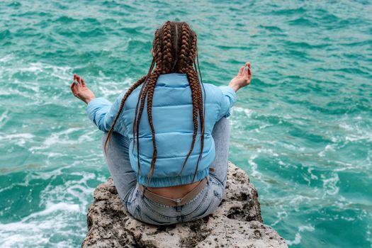 A woman in a blue jacket sits on a rock above a cliff above the sea, looking at the stormy ocean. Girl traveler rests, thinks, dreams, enjoys nature. Peace and calm landscape, windy weather