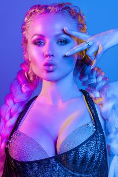 Fashion portrait of stunning sexy young blonde woman with trendy braids and sparkling bra under the net top. She is holding peace sign by the eye, making pouting lips in bright blue and red light